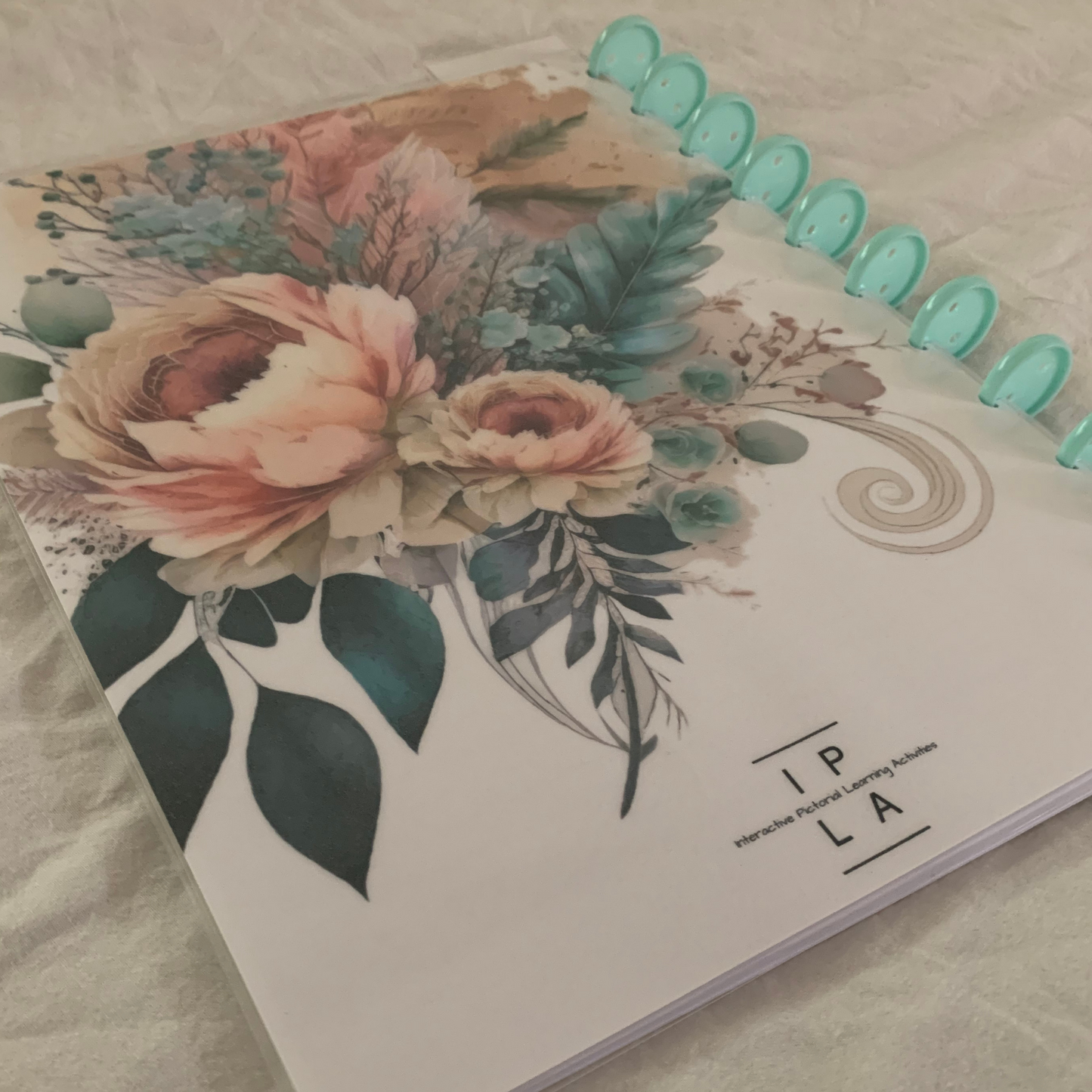IPLActivites logbook facilitates effective communication between parents and childcare staff. This picture shows the back of the logbook. A pastel watercolor flower covers the back just above the logo. 